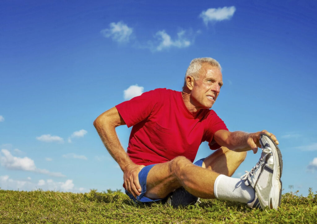 A male with a red t-shirt is sitting ont he ground & stretching out his hamstrings after a long run